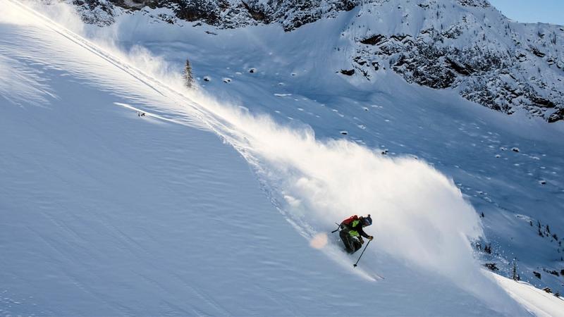 Skier making a sharp turn while descending a steep, snow-covered mountain slope, with snow spraying behind and rocky outcrops visible in the background.
