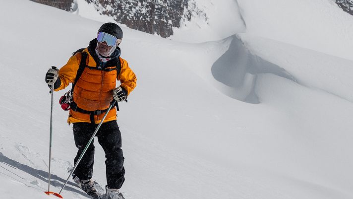 Person skiing on a snowy mountain slope, wearing a bright orange jacket, black helmet, and carrying ski poles.