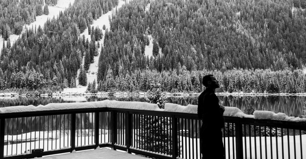 a black and white photo of a guest standing on a snowy balcony looking out at the vast tranquil tree-lined lake and mountains of the cmh heli-skiing area