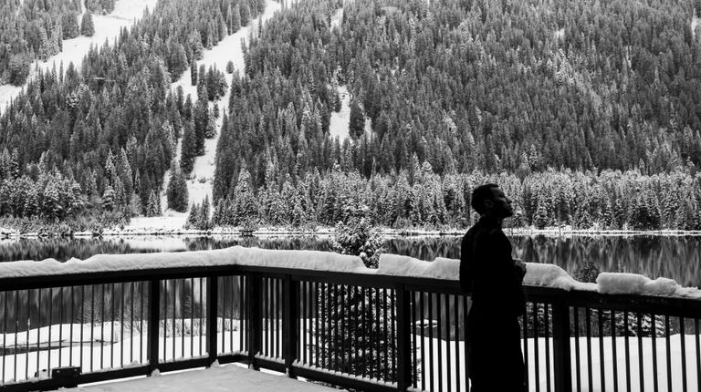 a black and white photo of a guest standing on a snowy balcony looking out at the vast tranquil tree-lined lake and mountains of the cmh heli-skiing area