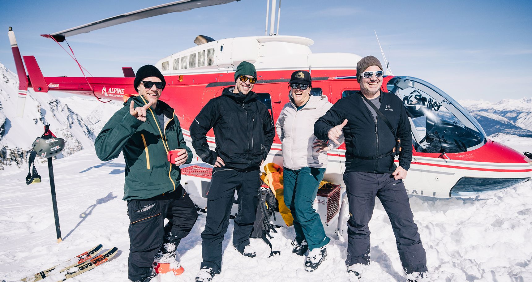 A small group of four skiers pose in front of a red CMH helicopter
