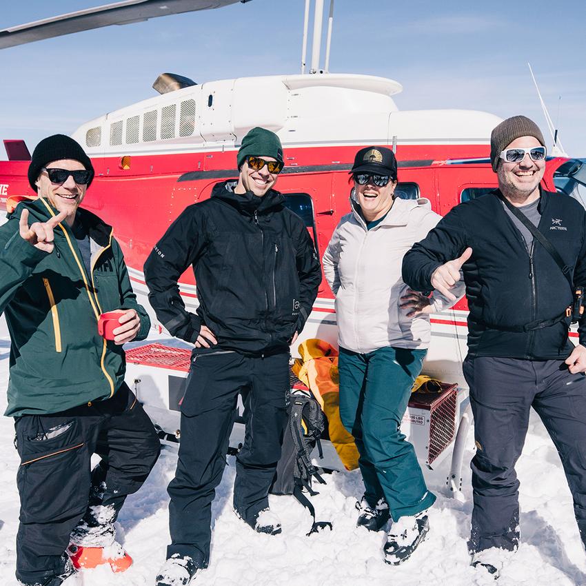 A small group of four skiers pose in front of a red CMH helicopter
