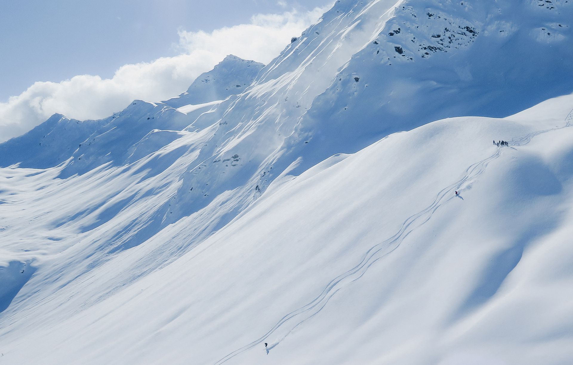Wide snowy slope with ski tracks and skiers descending under bright sunlight.