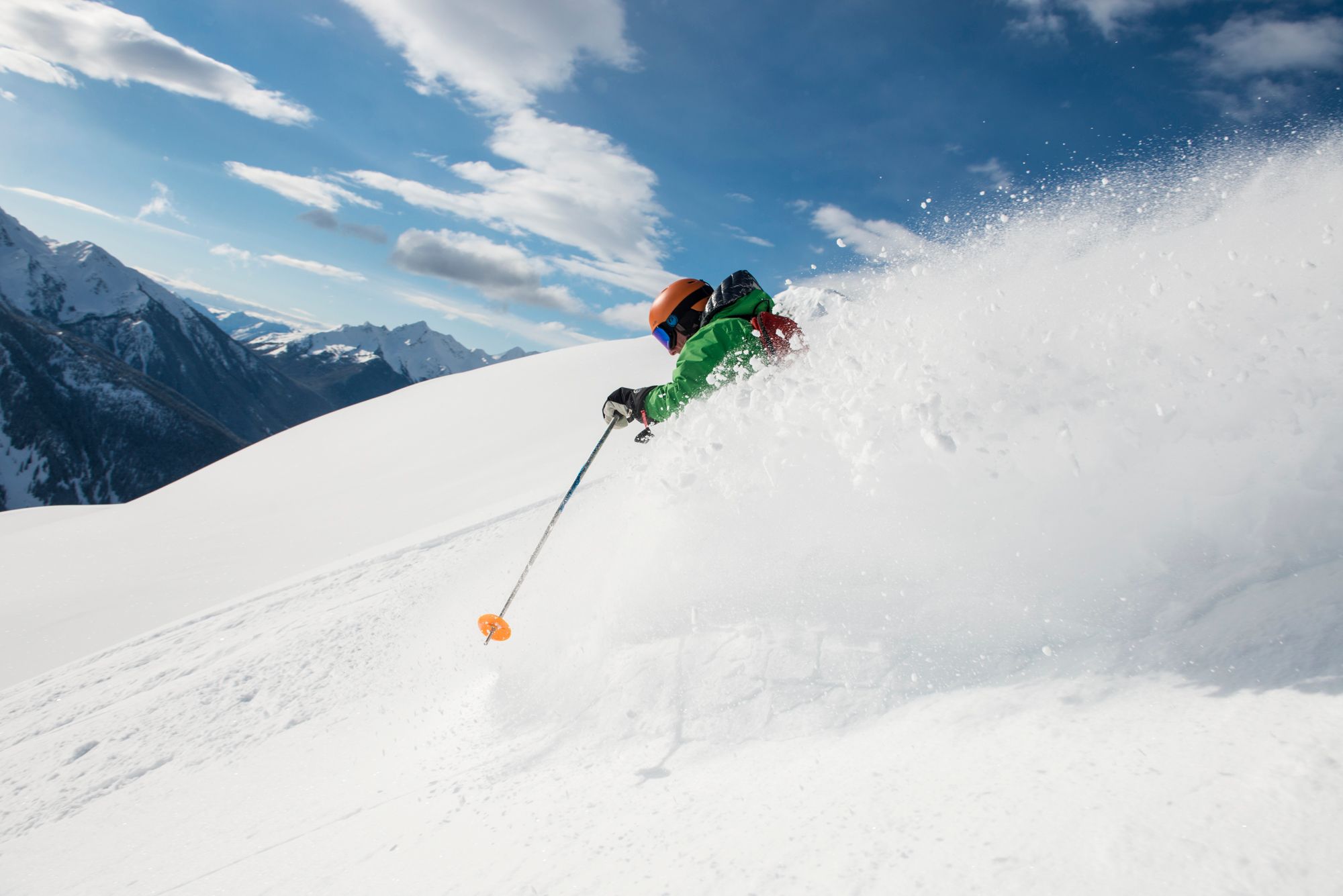 Skiier wearing a green coat and a road helmet makes their way down the mountain and sprays powder 