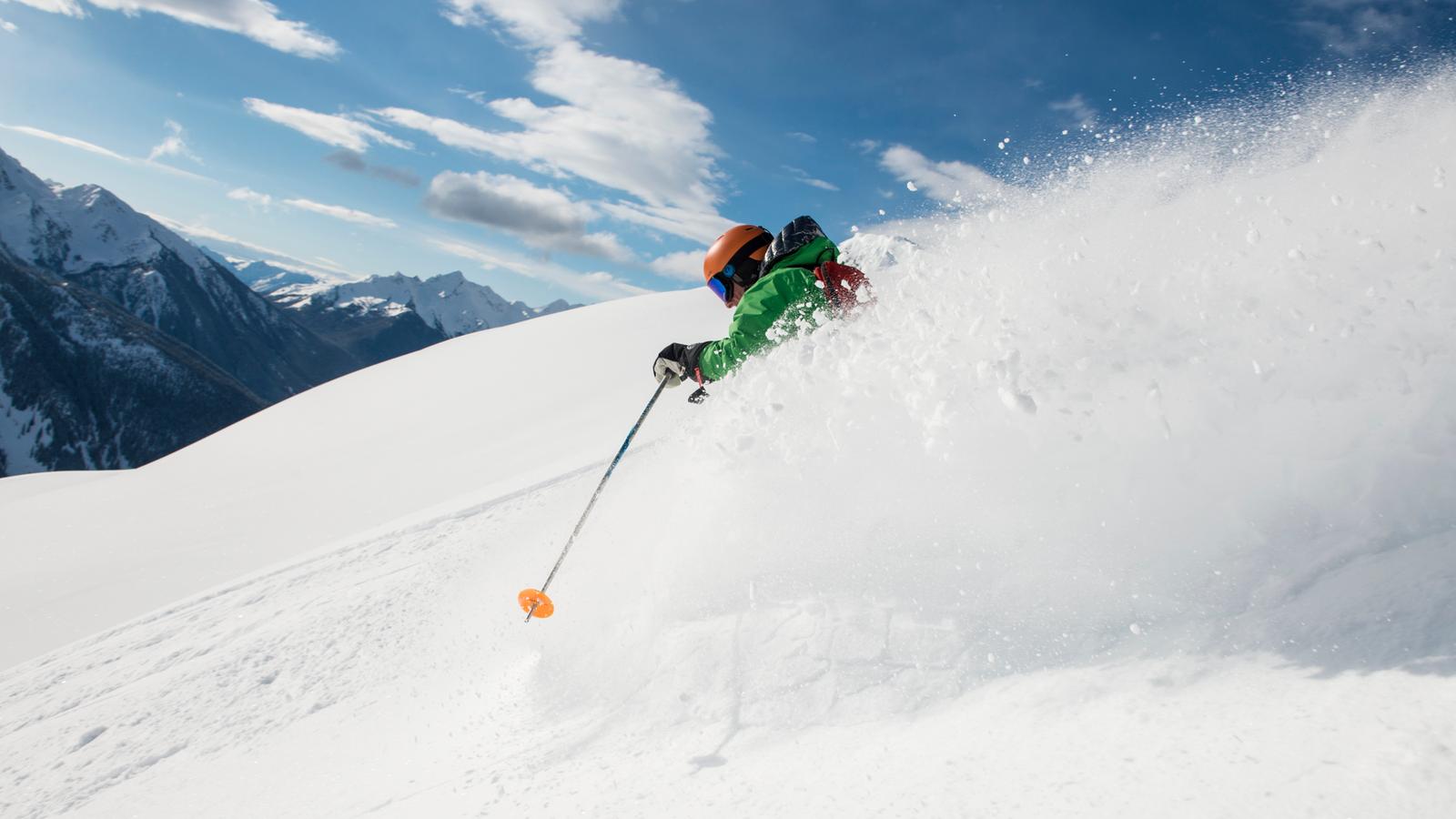Skiier wearing a green coat and a road helmet makes their way down the mountain and sprays powder