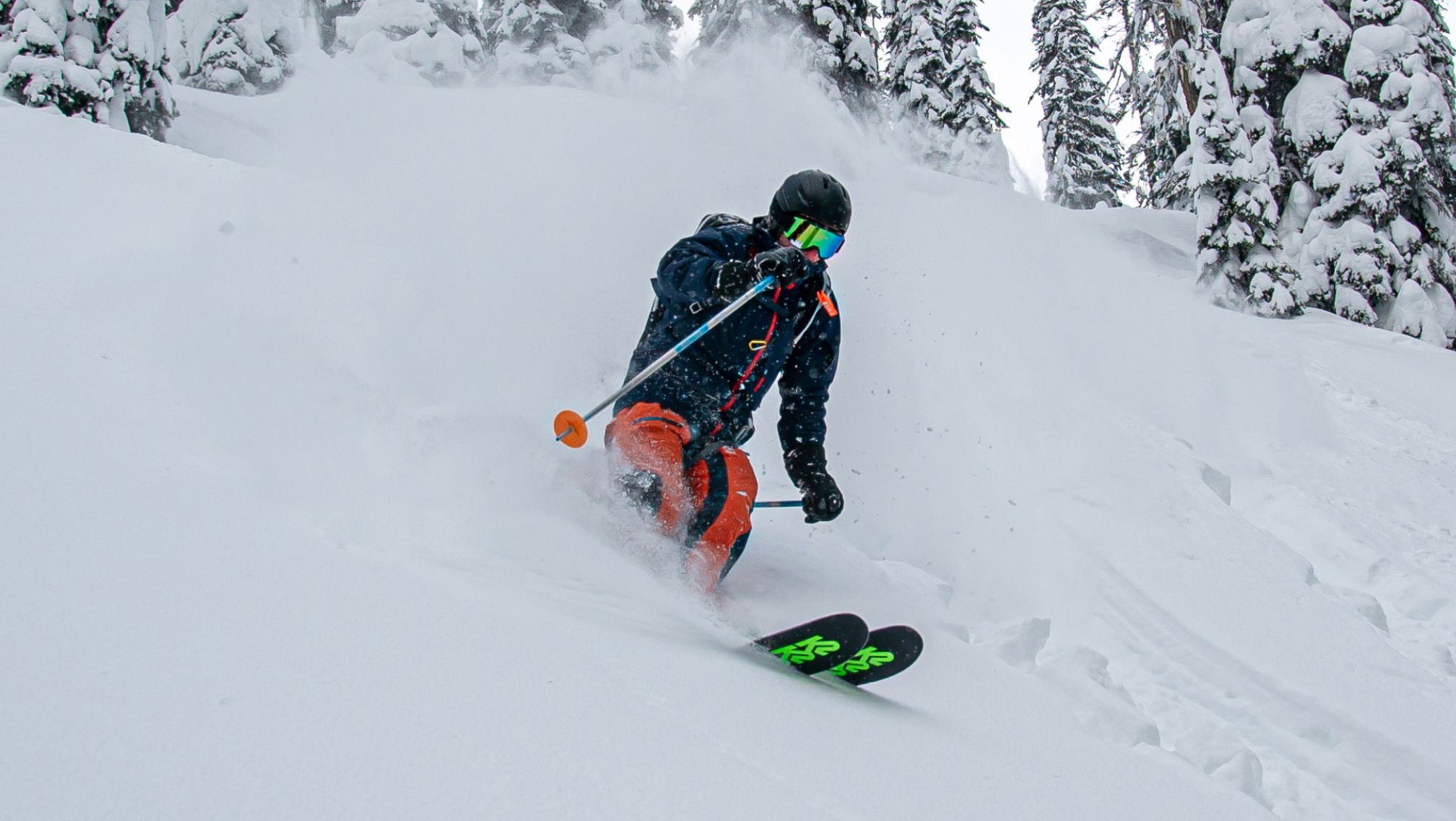 Skier in a dark helmet, reflective goggles, dark jacket, and bright orange pants skiing down a snowy slope, turning and spraying snow with snow-covered trees in the background.