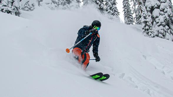 Skier in a dark helmet, reflective goggles, dark jacket, and bright orange pants skiing down a snowy slope, turning and spraying snow with snow-covered trees in the background.