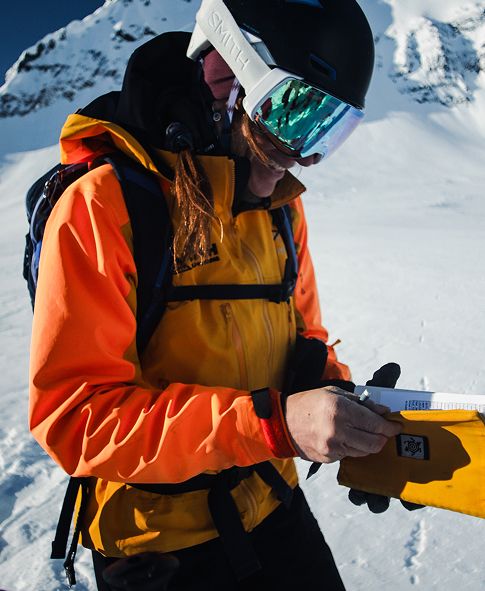 Person wearing a bright orange and yellow ski jacket and black helmet with reflective goggles, standing in a snowy mountain setting and holding a yellow notebook.