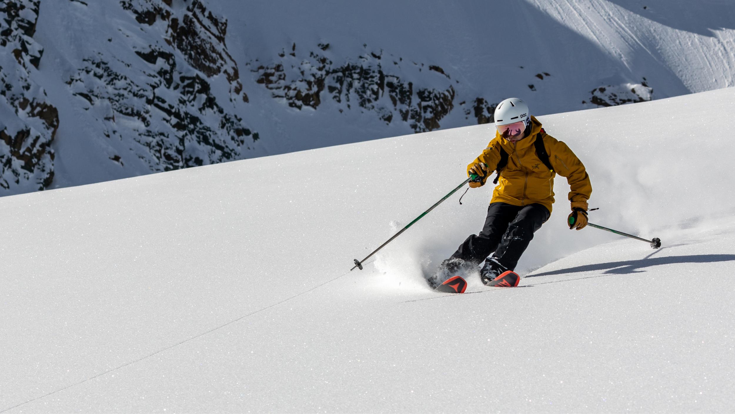 Skier wearing a rust yellow jacket and a white helmet carvs in some untouched snow powder