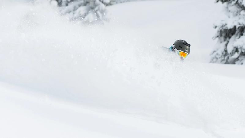 Person skiing or snowboarding through deep powder snow, wearing a helmet and goggles, with snow kicked up around them and snow-covered trees in the background.