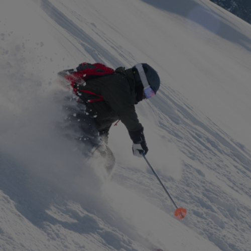 Skier carving through deep powder on a steep snowy slope, wearing a dark jacket, helmet, goggles, and a red backpack, with snow spraying up dramatically.