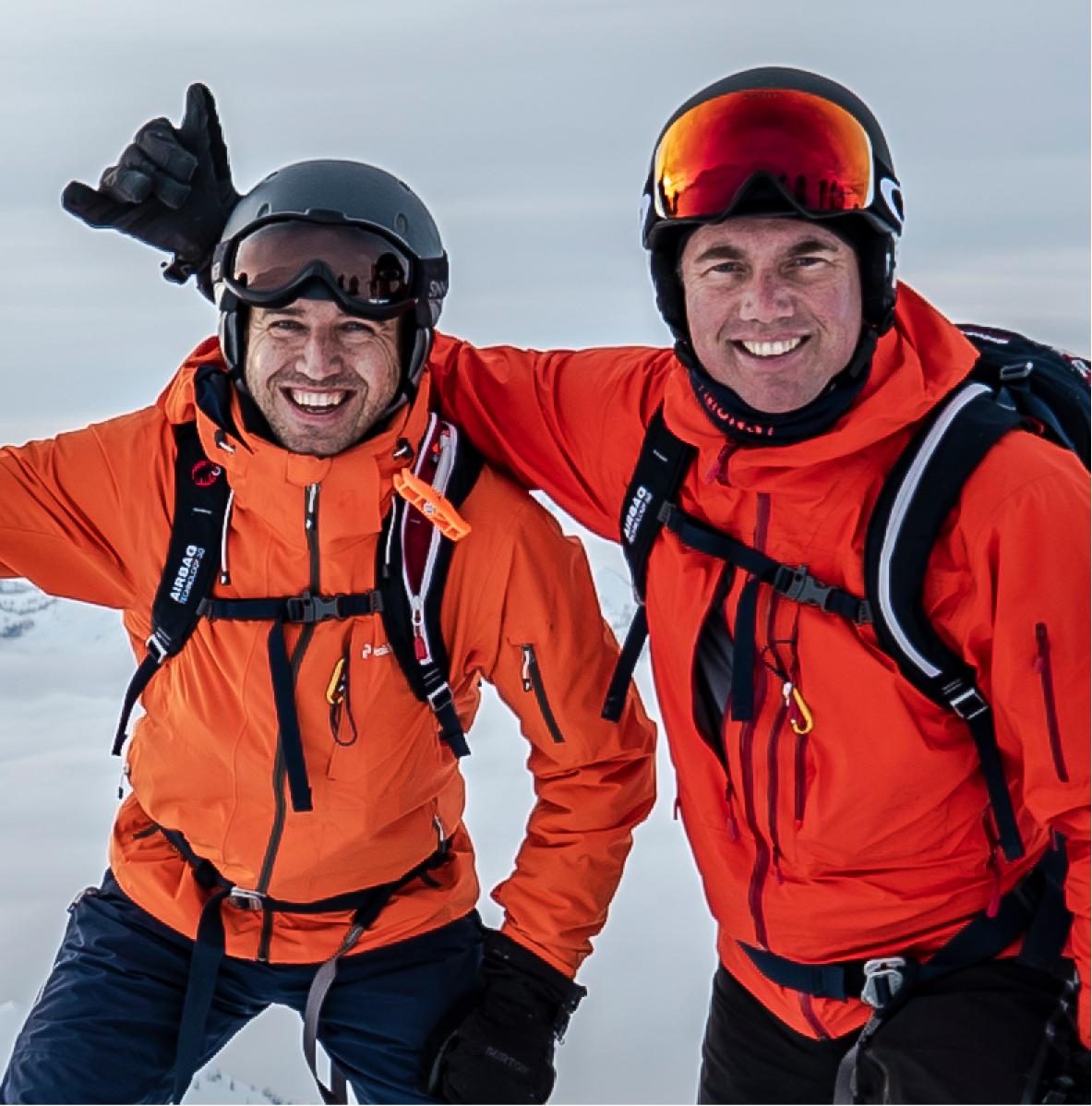 two skier skiers in orange jackets smile for a photo on a snowy mountaintop celebrating a successful day of cmh heli-skiing together