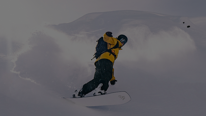 Person snowboarding down a slope in deep powder, wearing a bright yellow jacket and black helmet, with snow spraying up dramatically behind them against a backdrop of snowy mountains.