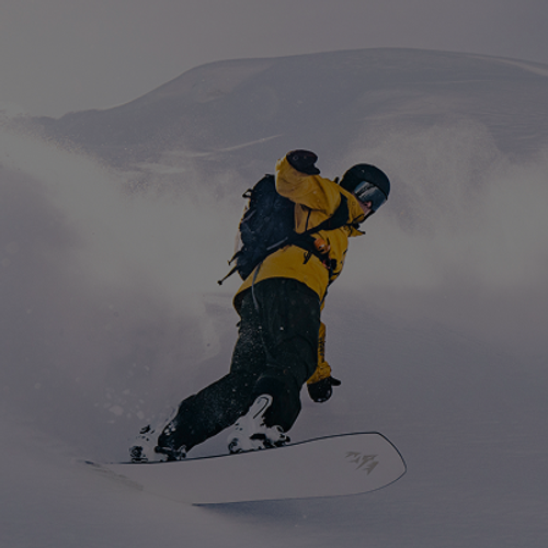 Person snowboarding down a slope in deep powder, wearing a bright yellow jacket and black helmet, with snow spraying up dramatically behind them against a backdrop of snowy mountains.