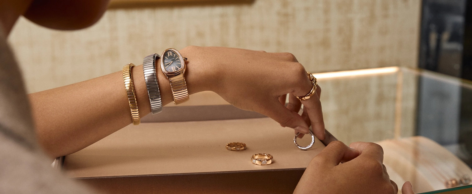 Gros plan d’une cliente essayant des bracelets de luxe et une montre en or et acier chez Château d’Ivoire, mettant en valeur des bijoux style Tubogas et des bagues en diamants dans une vitrine de boutique haut de gamme.