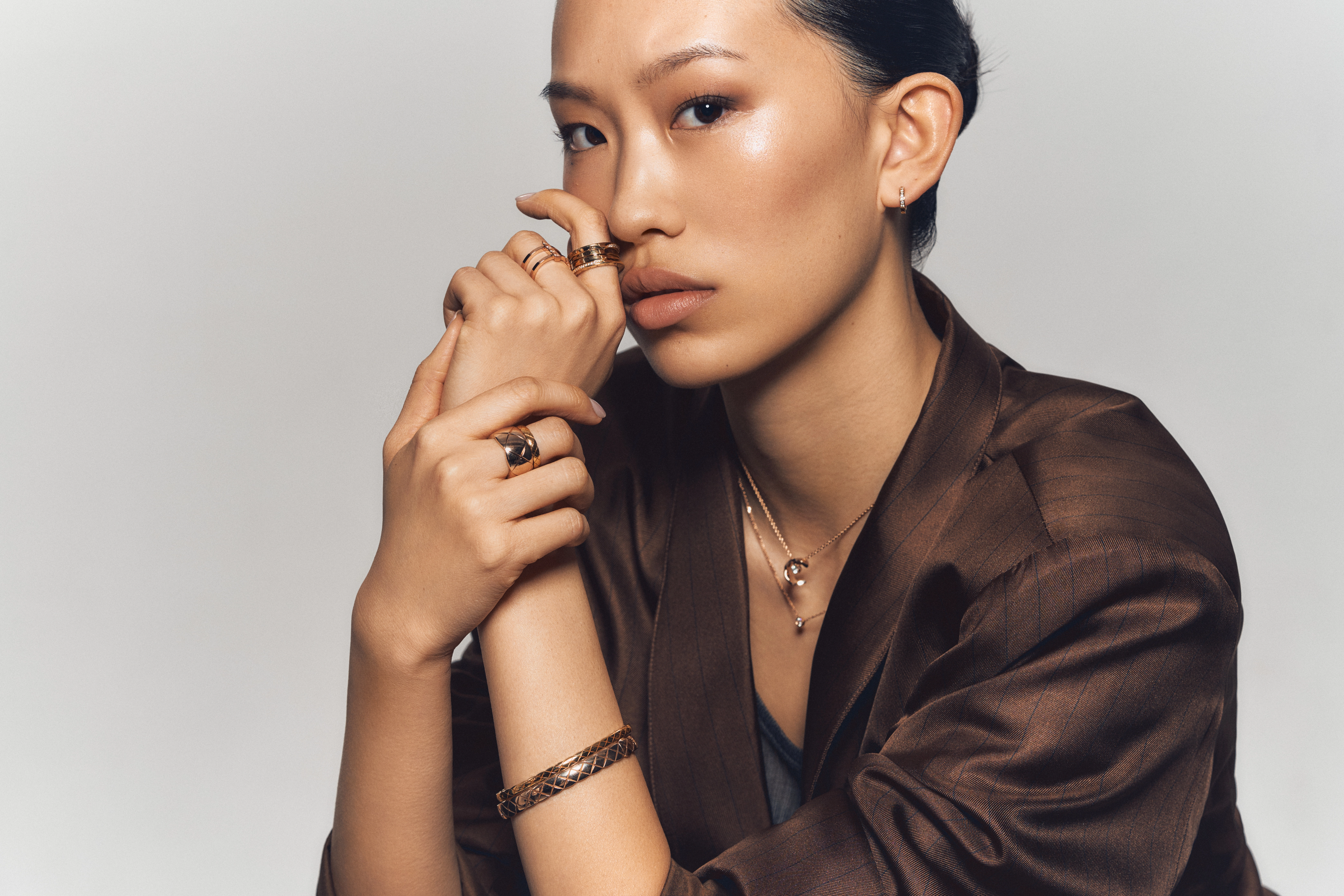 Close-up of a woman in a brown satin blazer wearing multiple gold rings, a quilted-pattern gold bracelet, and layered necklaces from the Chanel Coco Crush collection.
