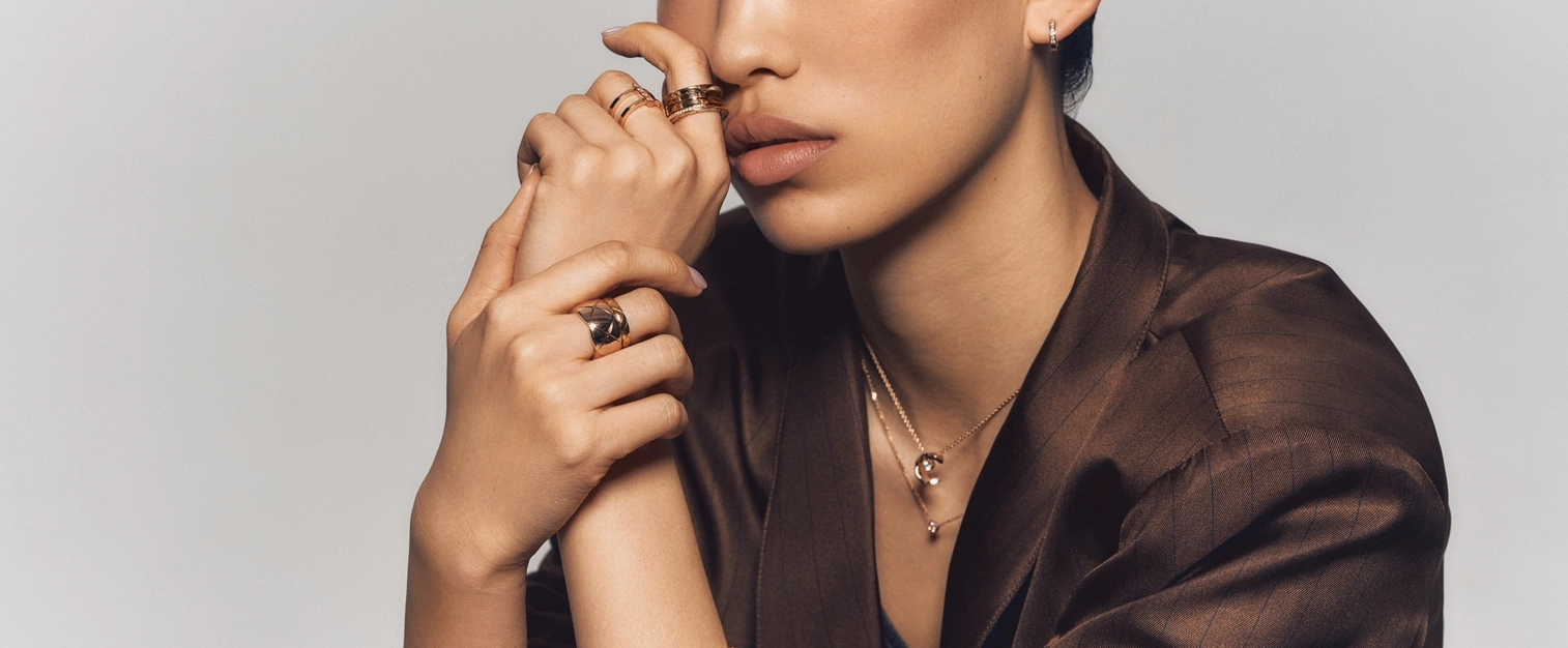 Close-up of a woman in a brown satin blazer wearing multiple gold rings, a quilted-pattern gold bracelet, and layered necklaces from the Chanel Coco Crush collection.