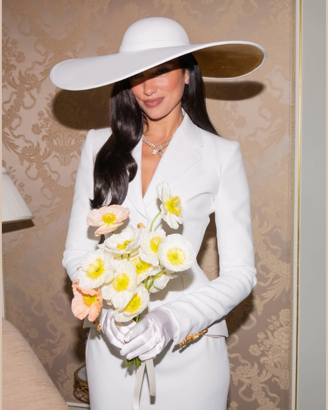 Dua Lipa portant un collier de Haute Joaillerie Bvlgari, un chapeau blanc et un tailleur blanc, tout en tenant un bouquet de fleurs.