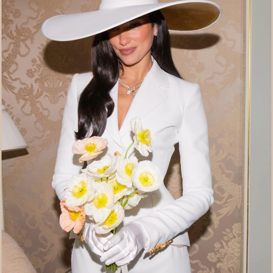 Dua Lipa portant un collier de Haute Joaillerie Bvlgari, un chapeau blanc et un tailleur blanc, tout en tenant un bouquet de fleurs.