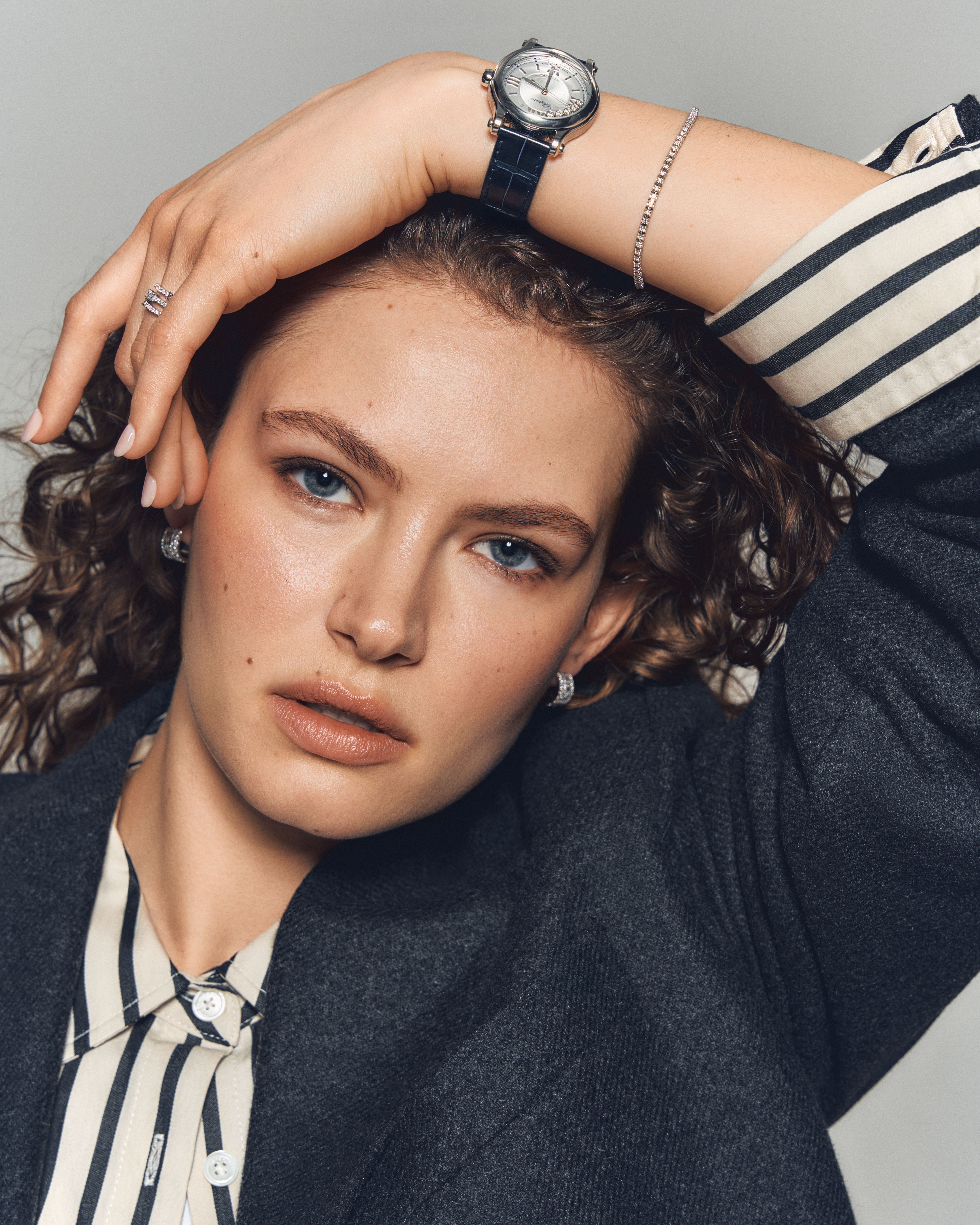 Close-up of a woman wearing a striped shirt and dark blazer, styled with a Chopard Happy Sport watch, a pavé diamond bracelet, a double-band diamond ring, and hoop earrings from Château D’Ivoire’s signature selection.