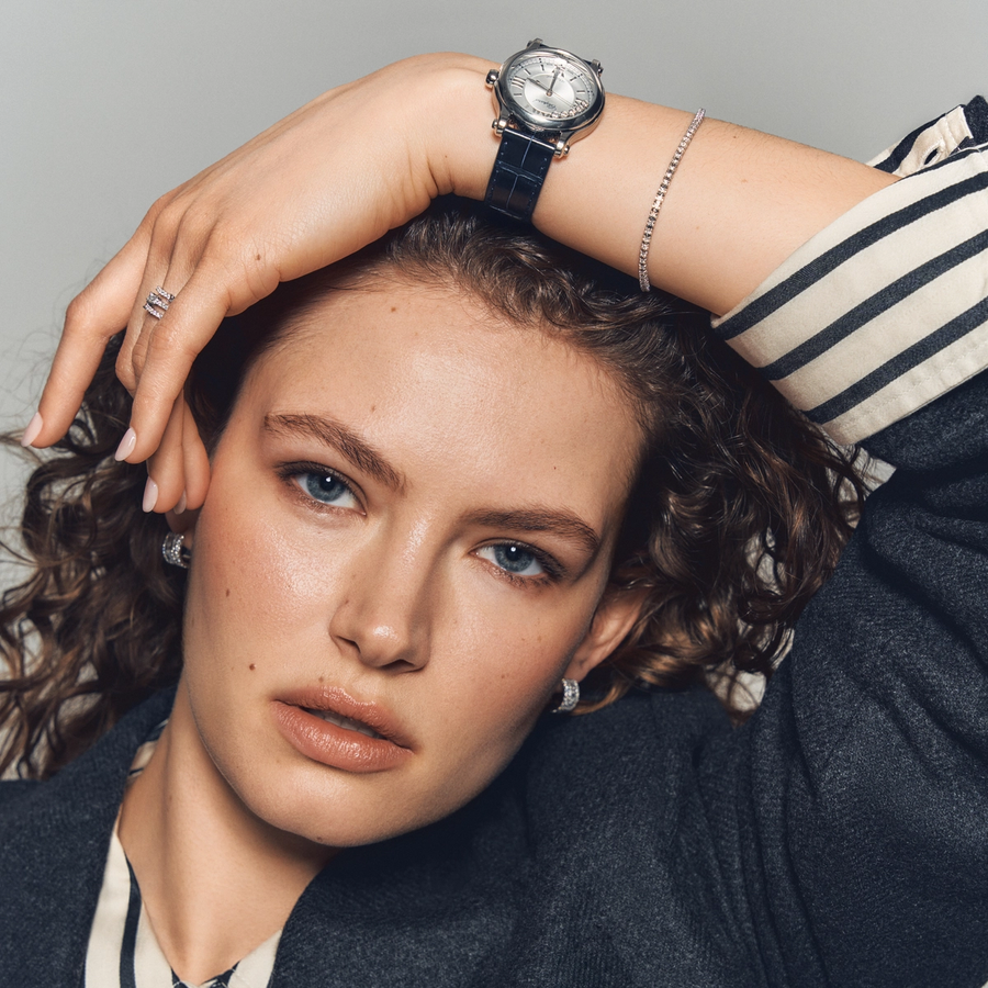 Close-up of a woman wearing a striped shirt and dark blazer, styled with a Chopard Happy Sport watch, a pavé diamond bracelet, a double-band diamond ring, and hoop earrings from Château D’Ivoire’s signature selection.