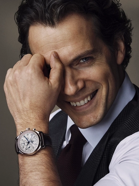 Portrait en gros plan d’Henry Cavill souriant, portant un gilet foncé sur une chemise blanche et une cravate bordeaux. Il couvre partiellement son visage avec sa main, mettant en valeur la montre Longines Flagship Heritage Moonphase à son poignet.