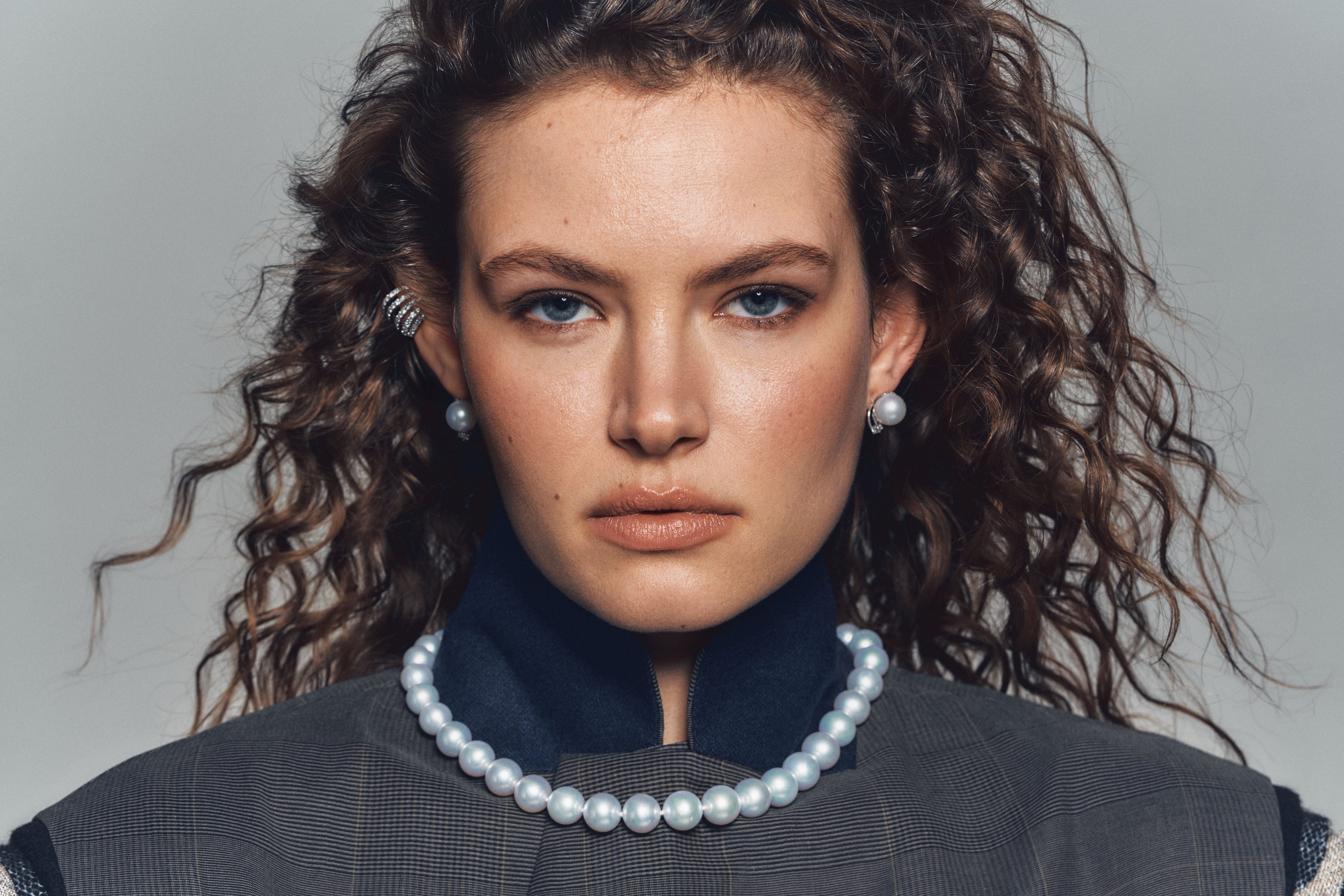 Close-up portrait of a woman with curly hair wearing a structured jacket, styled with luminous pearl earrings and a classic strand of round white pearls from Château D’Ivoire’s Perles du Château collection.