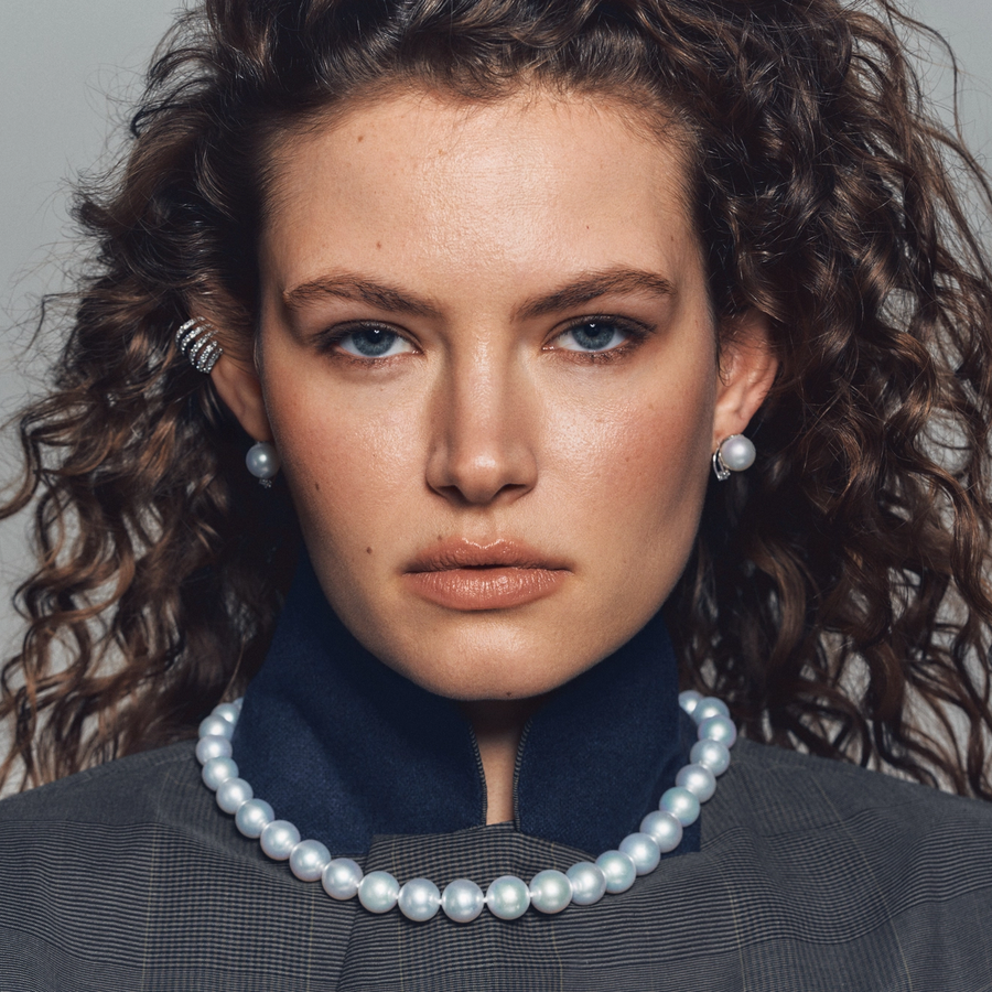 Close-up portrait of a woman with curly hair wearing a structured jacket, styled with luminous pearl earrings and a classic strand of round white pearls from Château D’Ivoire’s Perles du Château collection.