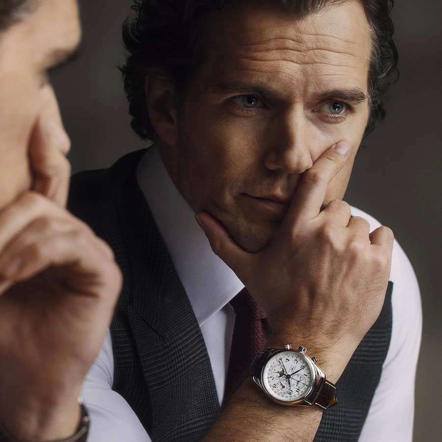 Henry Cavill regarde son reflet dans un miroir avec une expression pensive, la main posée contre son visage. Il porte un gilet à carreaux foncé sur une chemise blanche et une cravate bordeaux, mettant en valeur la montre Longines Flagship Heritage Moonphase à son poignet.