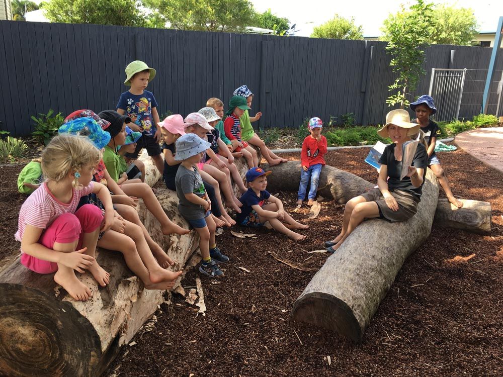 Children gathered on log, reading a book with a teacher