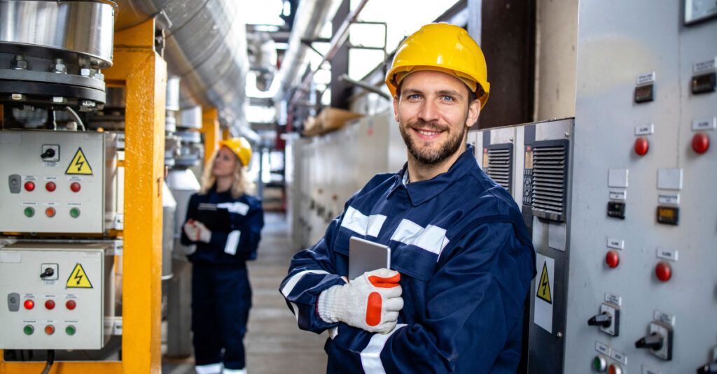 A man in a blue work uniform and a yellow hard hat stands confidently in an industrial setting. He is smiling and holding a smartphone in one hand, while the other hand is resting on his arm. Behind him, there are control panels with various buttons and indicators, and a woman in a similar uniform is seen in the background, looking at a clipboard. The environment features pipes and machinery, indicating a factory or processing plant.