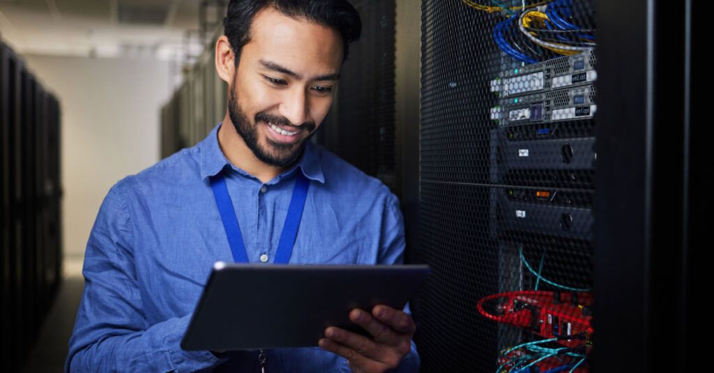 A man with a beard is smiling while holding a tablet in a server room. He is wearing a blue shirt and a blue lanyard. Behind him, there are server racks with various cables and equipment visible, including network switches and servers. The environment is well-lit, emphasizing the technology around him.