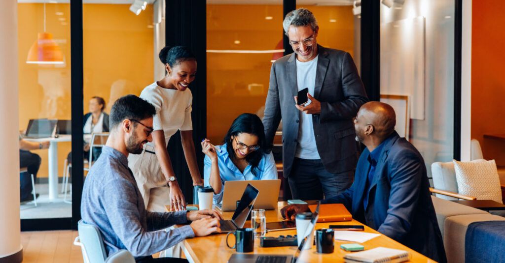 A group of five professionals is engaged in a collaborative meeting around a wooden table in a modern office setting. Two individuals are seated, one working on a laptop and the other looking at a notebook. Three others are standing, with one pointing at the laptop screen while another smiles and holds a coffee cup. The atmosphere is lively and focused, with bright orange walls and contemporary furnishings in the background.