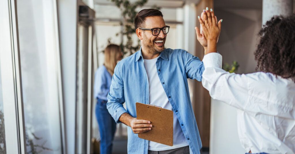 A man in a light blue shirt and glasses is smiling and giving a high five to a woman wearing a white shirt. He holds a clipboard in his left hand. In the background, another person in a blue shirt is partially visible, standing near large windows that let in natural light. The setting appears to be a modern office environment.