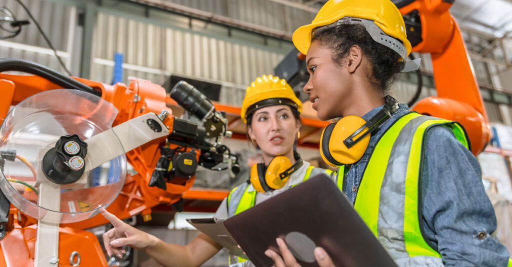 Two women in safety gear are engaged in a discussion near an industrial robotic arm. Both are wearing yellow hard hats and high-visibility vests. One woman is pointing at a control panel with gauges, while the other is holding a tablet and listening attentively. The background features a warehouse setting with shelves and machinery.