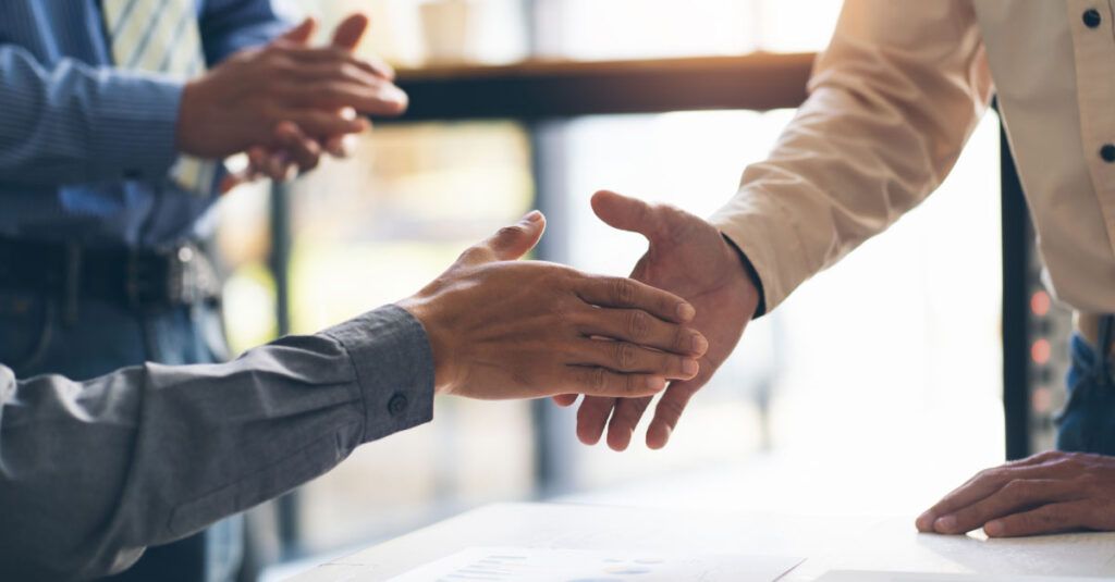 A close-up of two hands engaged in a handshake, symbolizing a business agreement or greeting. One hand is wearing a gray long-sleeve shirt, while the other is in a light-colored shirt. In the background, there are blurred figures and a bright, well-lit environment, suggesting a professional setting. A document with charts is visible on the table beneath the hands.