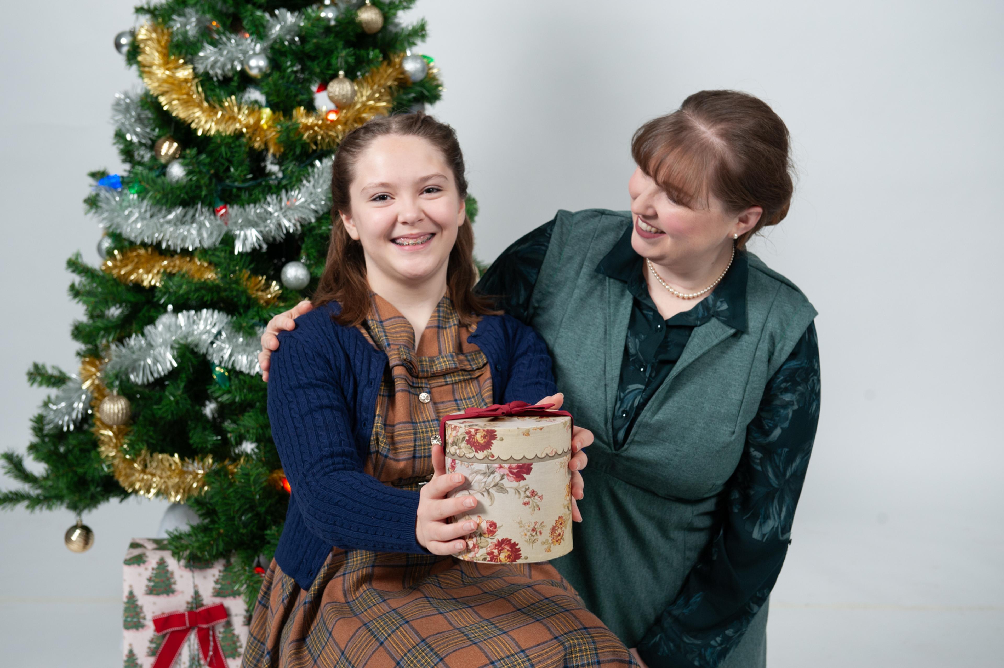 A young girl holding a present as her mother looks on with a Christmas tree in the background