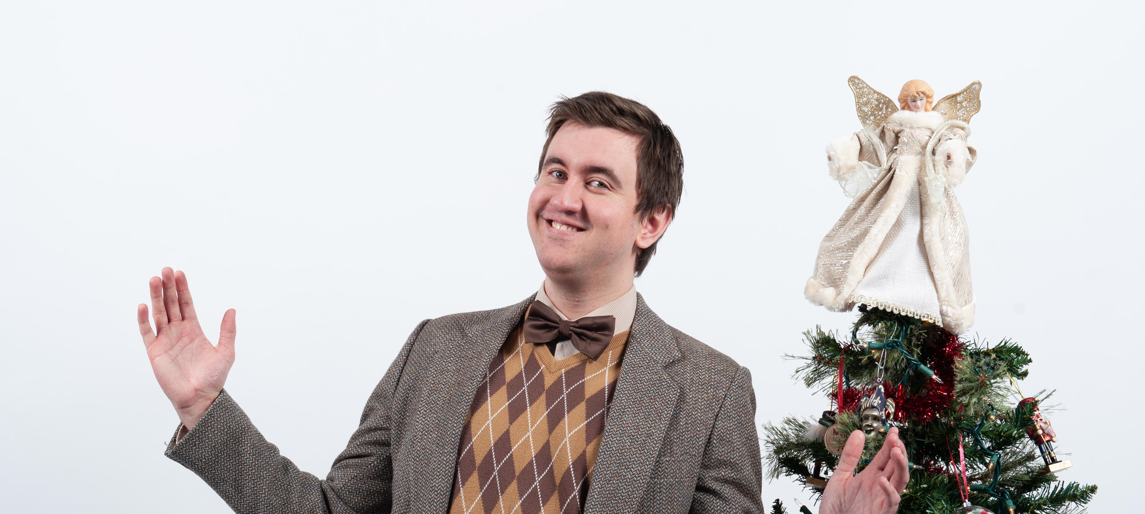 A man with his arms open wide, standing in from of a decorated Christmas tree.