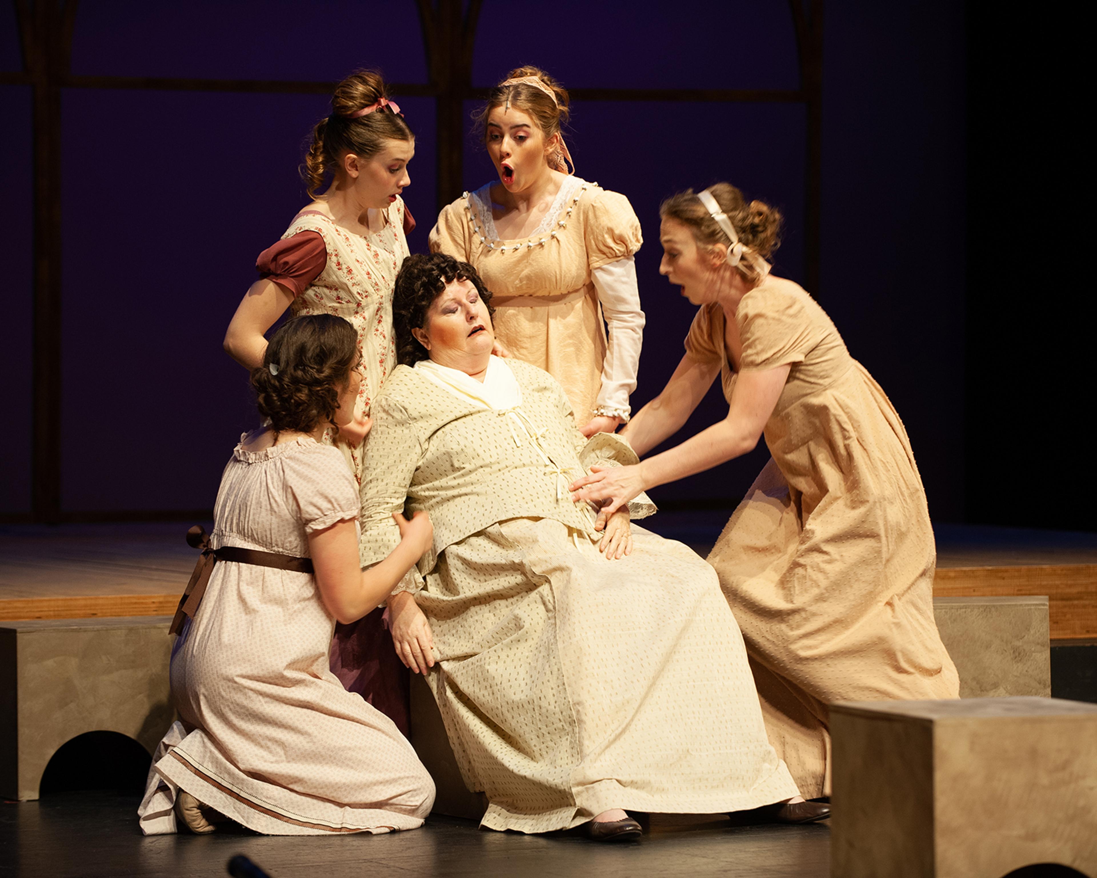 A production photo from Pride and Prejudice with Mrs. Bennet fainting into a chair. Her daughters, who are gathered around her, look shocked.