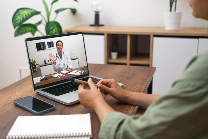 A person at a wooden desk has a video call with a smiling clinician on a laptop, from a sunlit room at home.