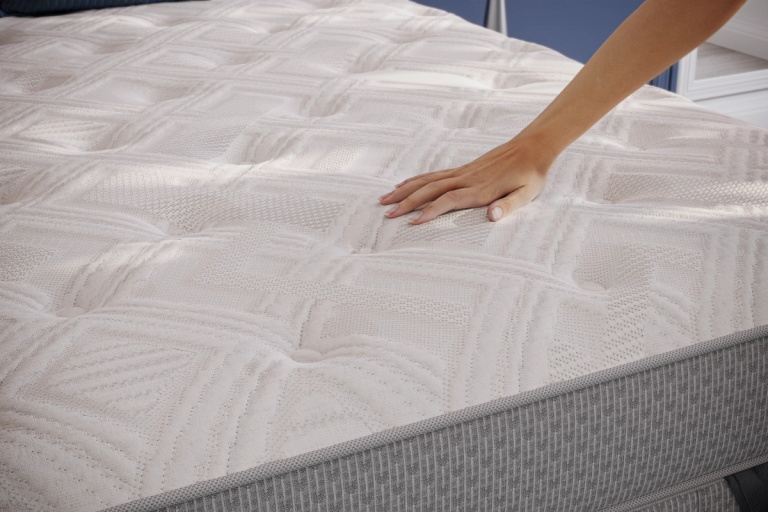 A hand touches a white quilted mattress with a grey side.