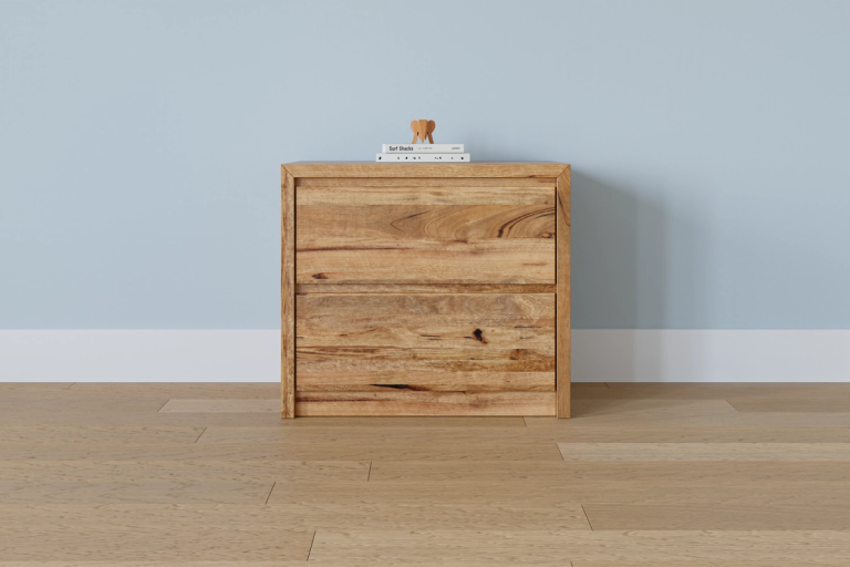 A two-drawer natural wood nightstand with books and a wooden elephant on top, against a light blue wall and wood floor.