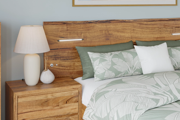 A modern bedroom with a wooden headboard, wooden nightstand, a white lamp, and green and white tropical leaf-patterned bedding.