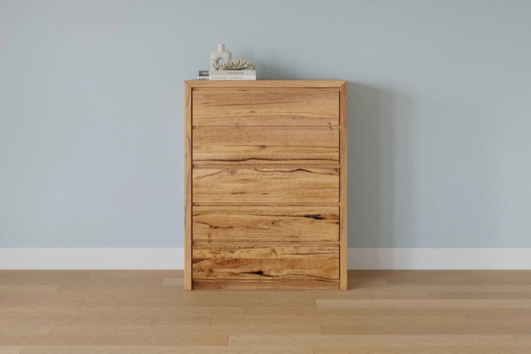 A natural wood five-drawer dresser stands against a light blue wall and hardwood floor, topped with books and a decorative object.