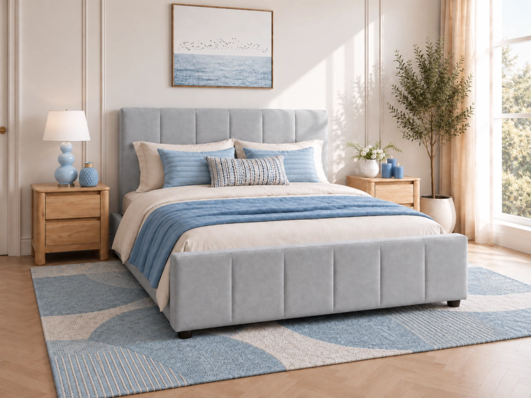 A serene bedroom featuring a light blue upholstered bed with beige bedding and blue accents, flanked by wooden nightstands, on a blue and beige patterned rug. An ocean art print hangs above the bed.