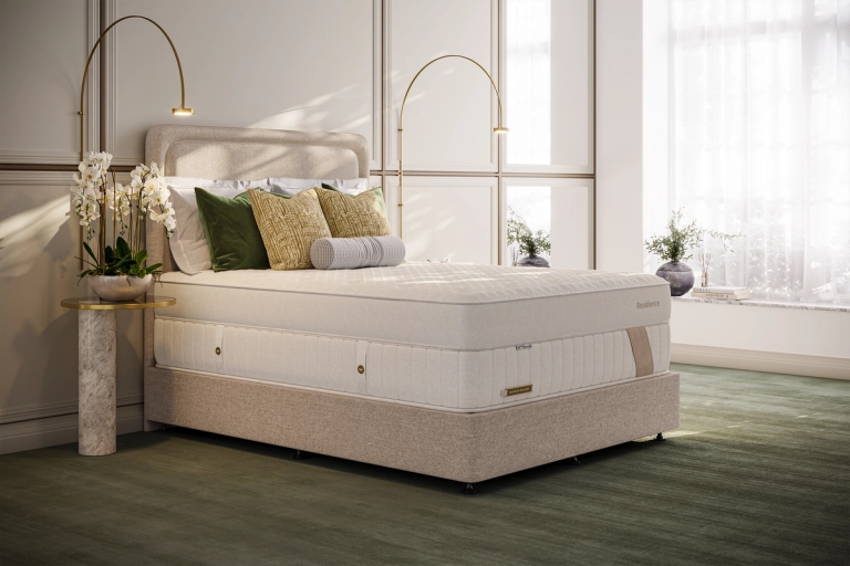A modern bedroom features a beige bed with a thick cream mattress, decorative pillows, two tall brass lamps, and an orchid on a marble side table.