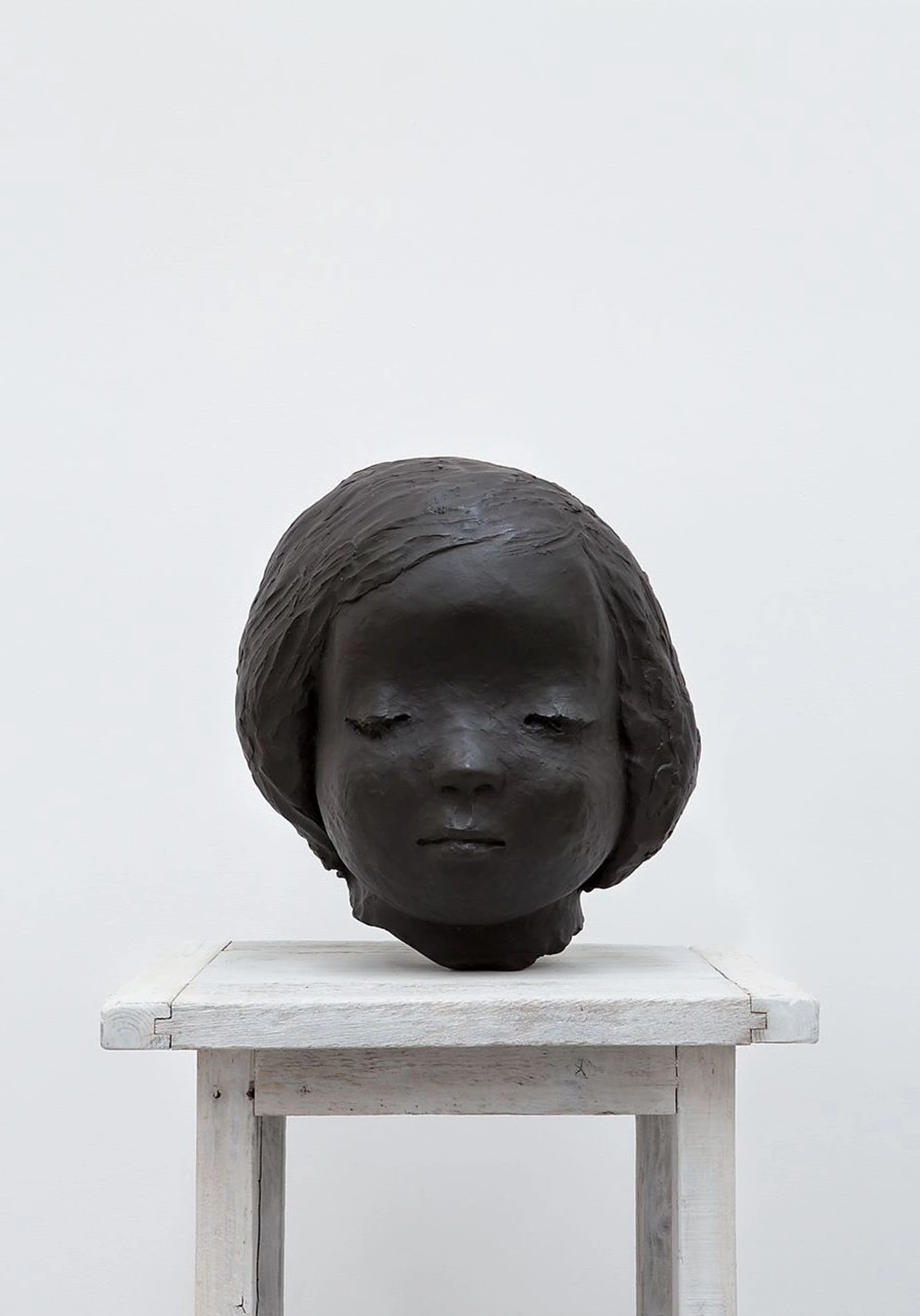 Bronze head sculpture of a girl with closed eyes and a bob, displayed on a white painted timber bench.