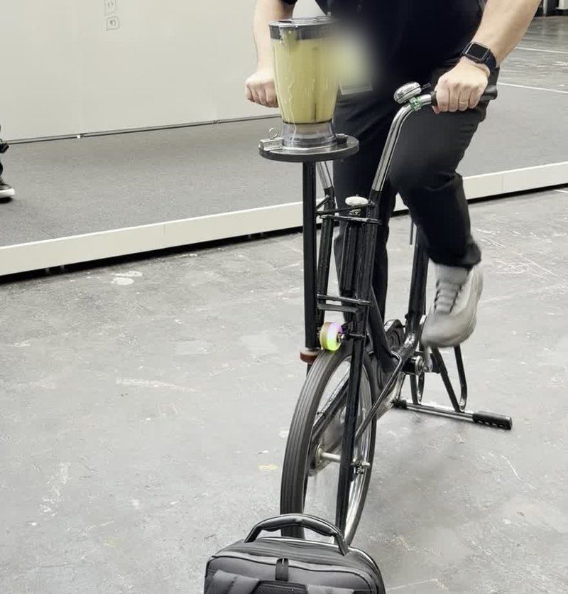 A bicycle on a stand with a smoothie blender on a post in front of the handlebars, a rider pedalling, seen from the chest down, on the grey expo floor