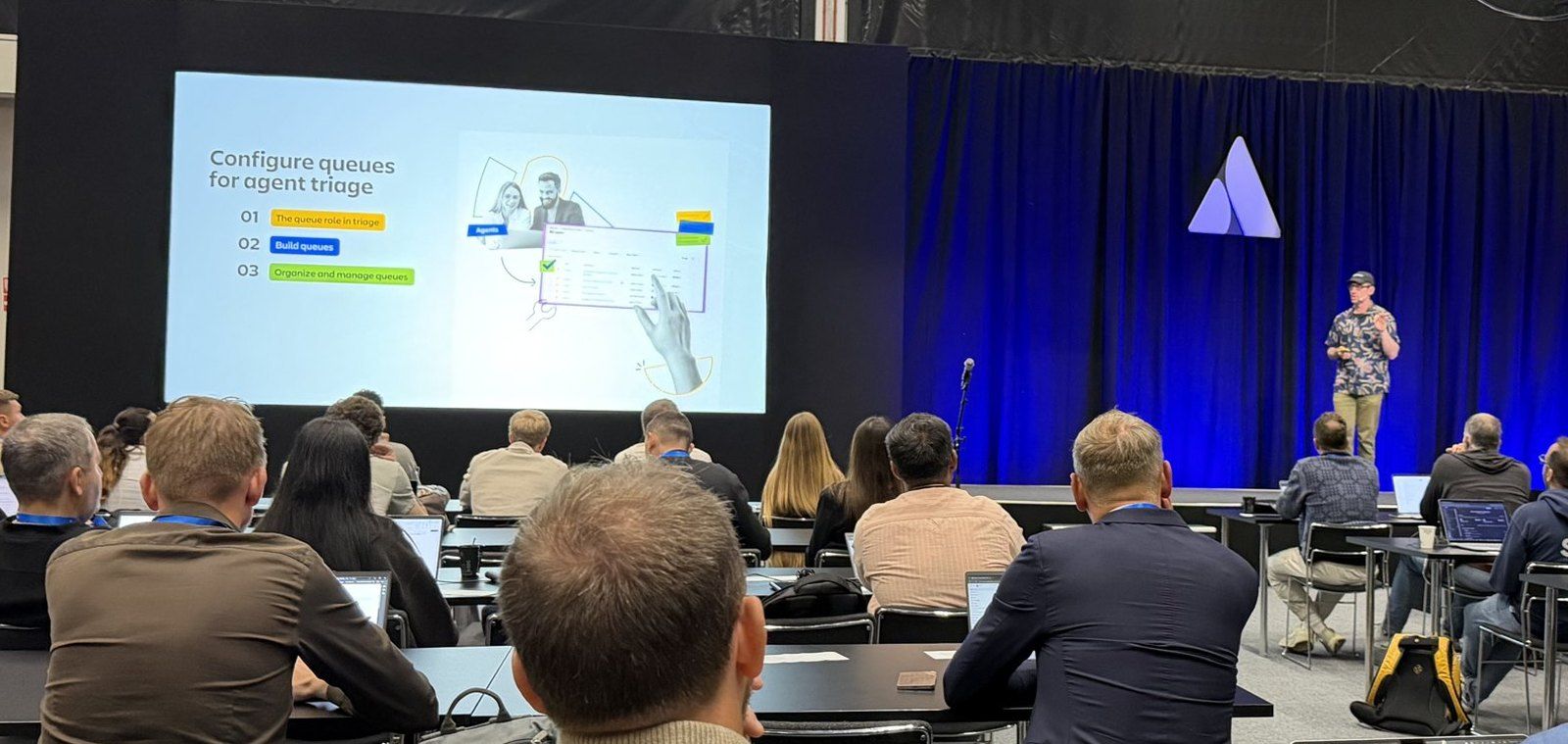 A breakout hall seen from the back rows: a large slide reading Configure queues for agent triage with the agenda 01 The queue role in triage, 02 Build queues, 03 Organize and manage queues, a speaker on a blue-curtained stage, and attendees at long tables with laptops