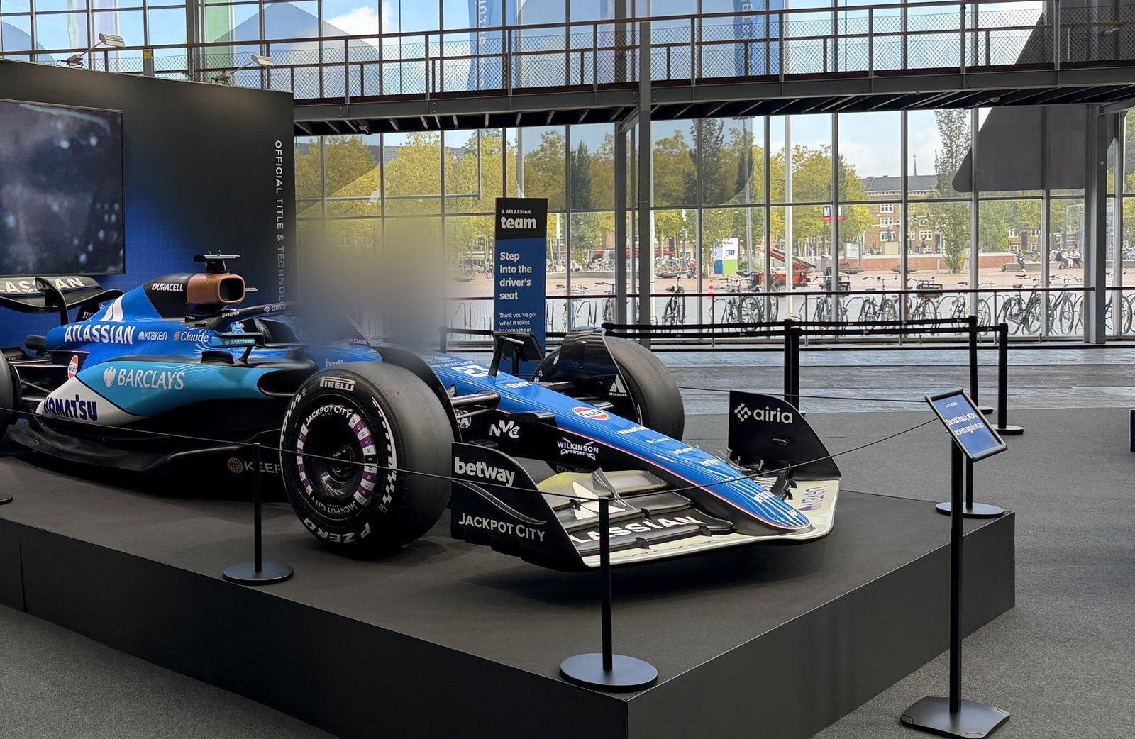 An Atlassian Williams F1 Team show car in blue livery on a black plinth inside a glass-walled hall, with vertical backdrop text reading OFFICIAL TITLE & TECHNOL, the rest hidden behind the car, and a sign reading Step into the driver's seat